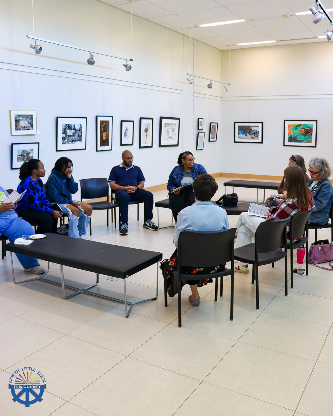 A group of people sit in a circle in an art gallery, participating in an interactive story circle.
