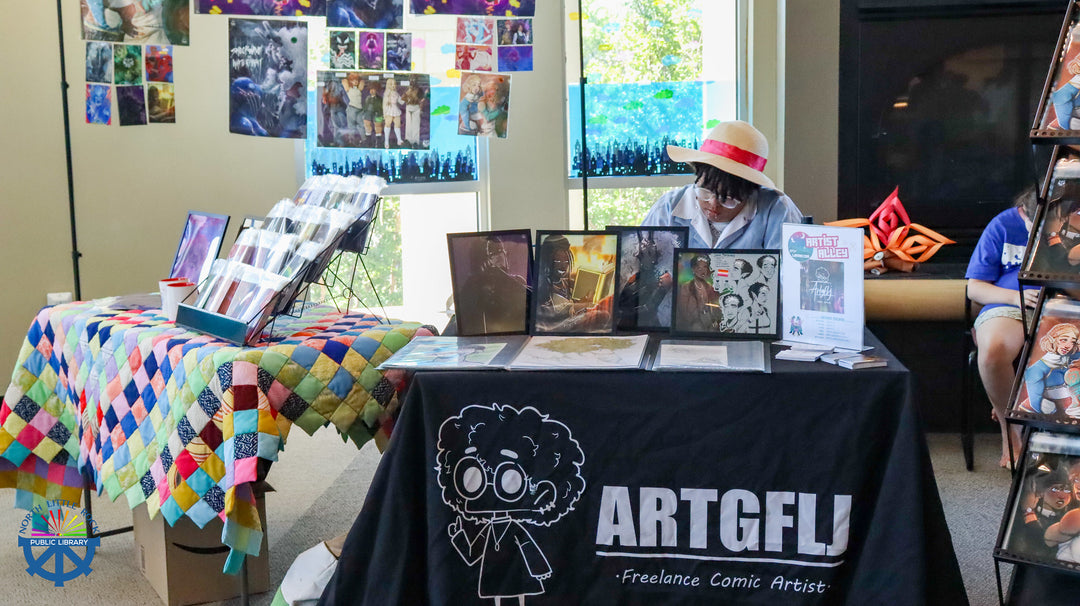 Laman Con Artist drawing comic and anime style characters in their Art booth with 'ARTGFL' tablecloth featuring a cartoon character, various art pieces, and promotional materials.