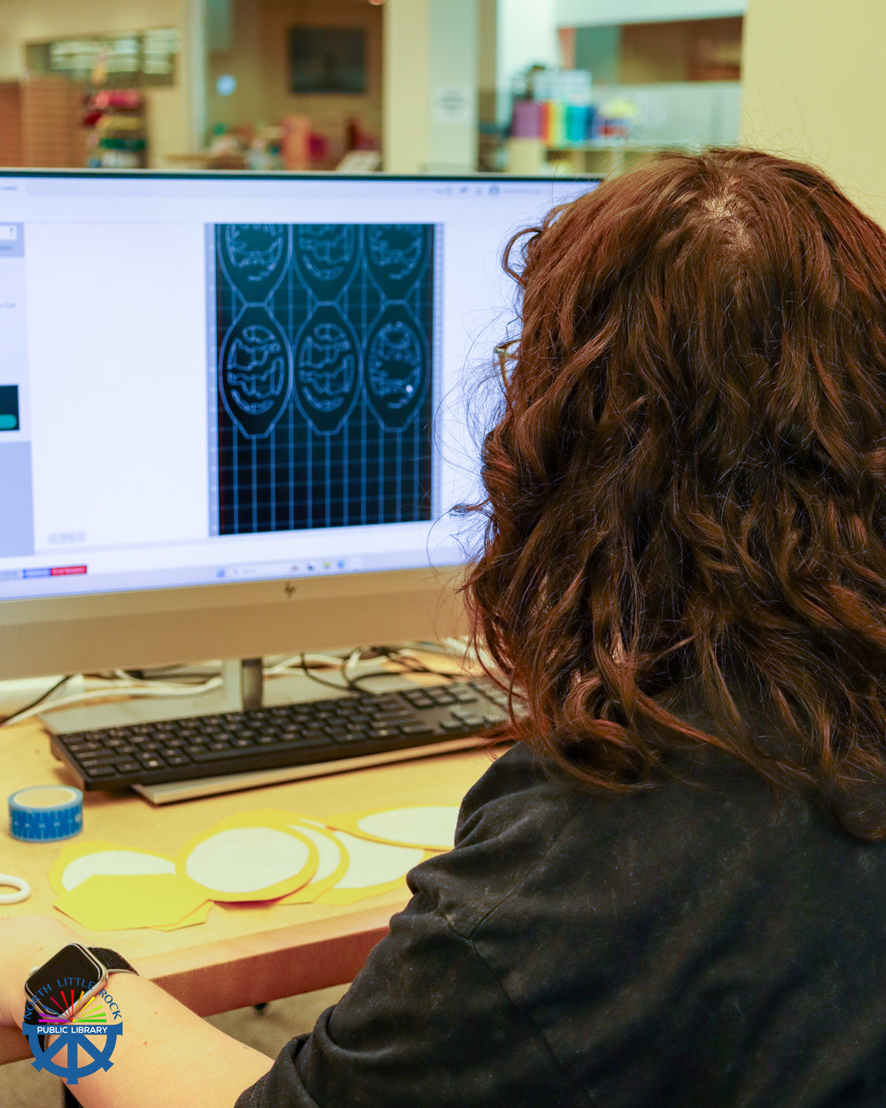 A person sitting at a computer with an image on the screen. Cut pieces in that image are on the table in front of the computer.