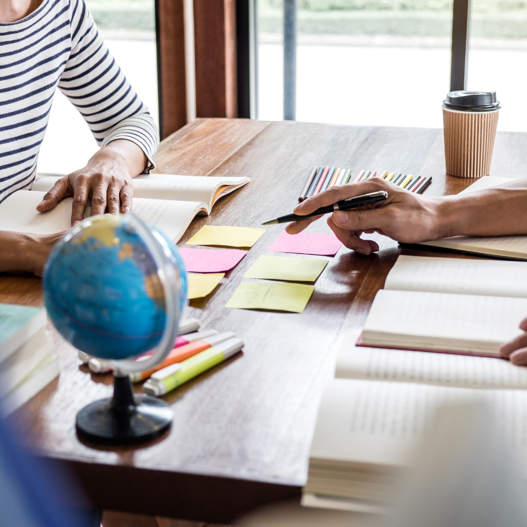 Two people sitting at a wooden table with open books, sticky notes, markers, and pencils, with a coffee cup in the background and a globe in the foreground.