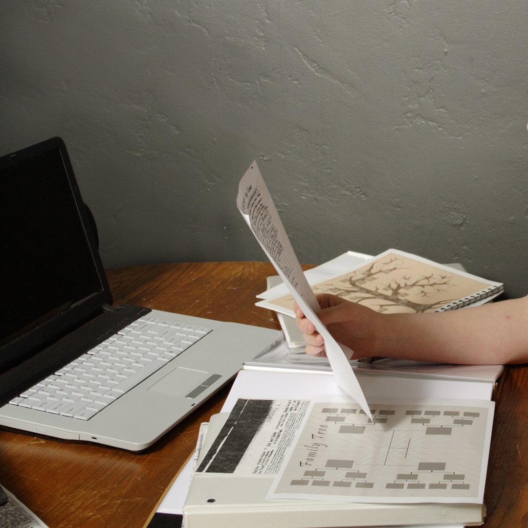 A table with a laptop and various papers and documents pertaining to genealogical research. A hand holds up one document.