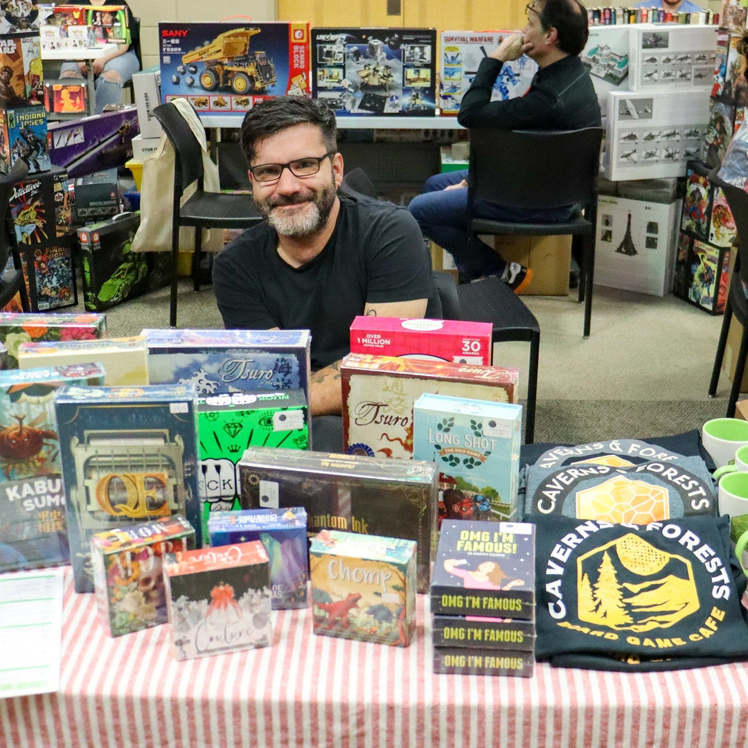 Caverns and Forests owner sitting at a table with board games and merchandise on display for sale.