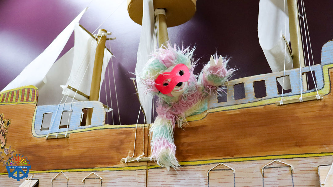 Plush toy with a pink superhero comic mask on a wooden sailboat against a purple background