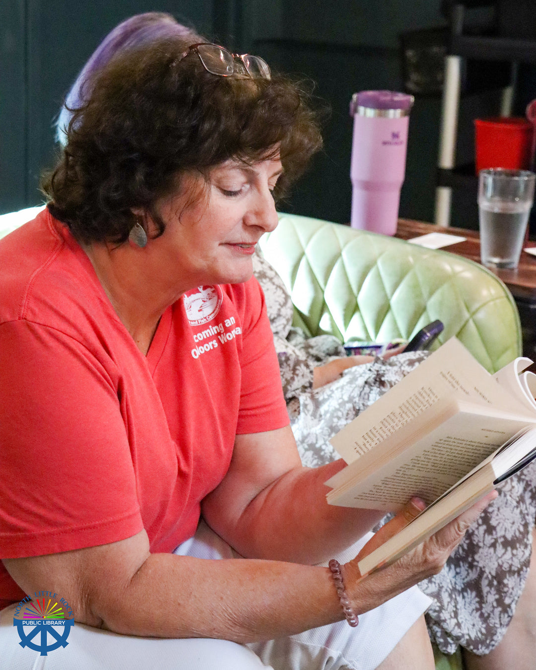 Woman in a red shirt reading the one community book in a casual setting with a green chair and pink cup in the background.