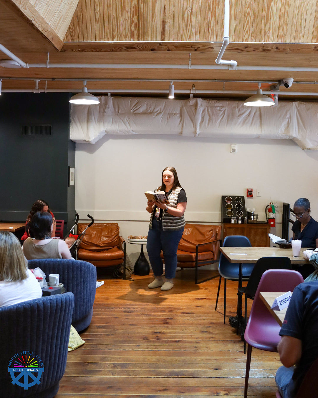 Woman giving reading from a book to an audience in a casual setting with chairs and tables.