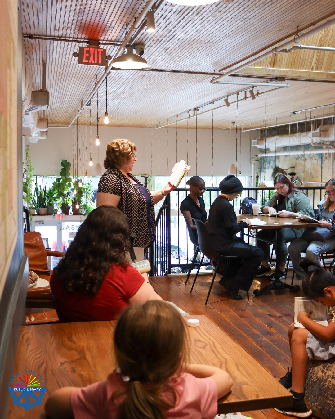 People sitting at tables reading along with Megan in a modern cafe with large windows.