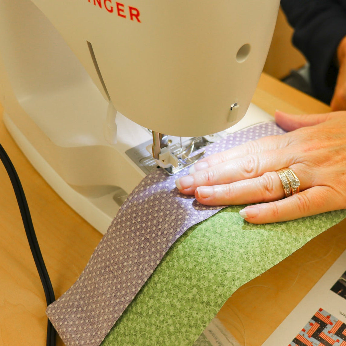 A close-up of a Singer sewing machine being used to sew together purple and green fabrics.