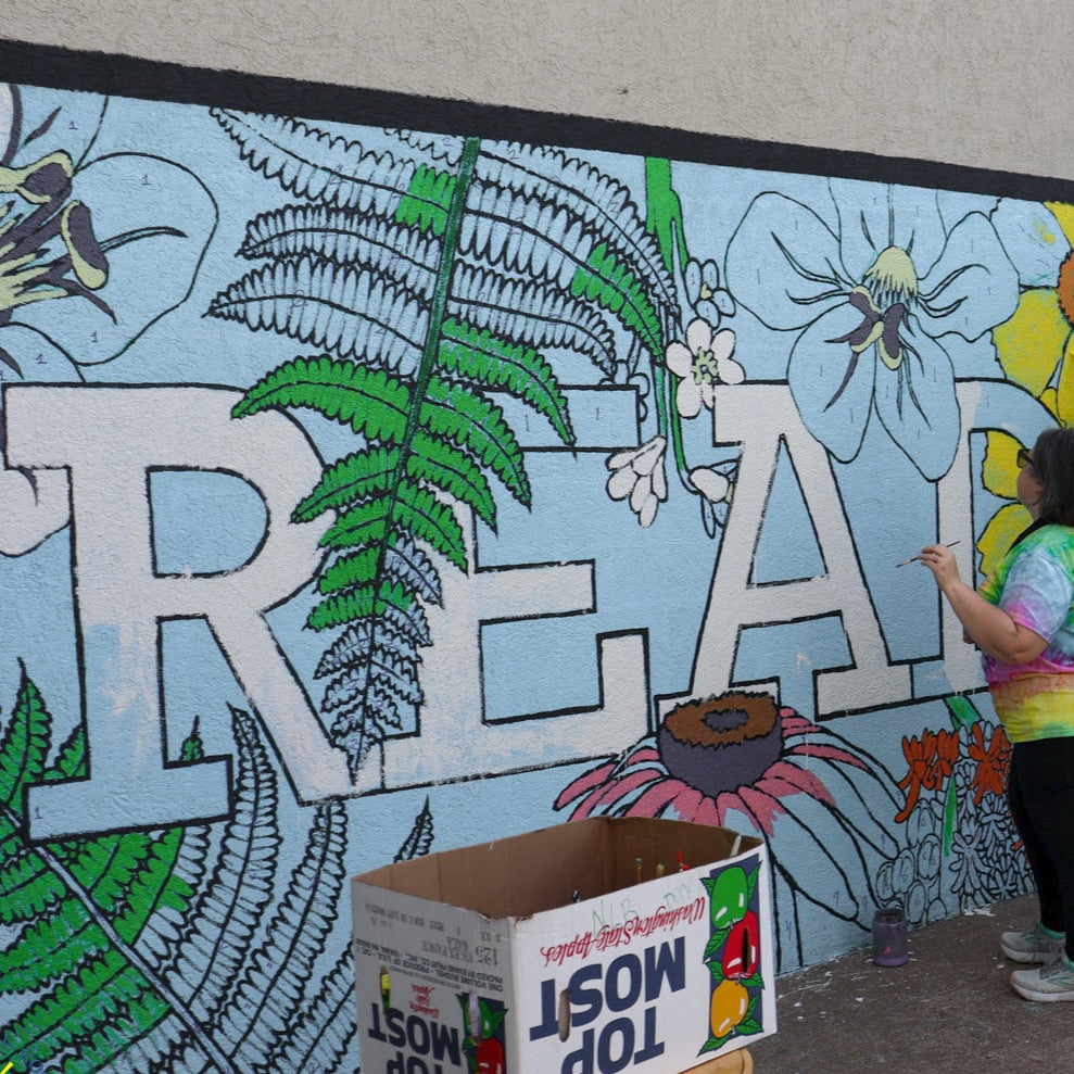 Two people painting a large mural of flowers and the work Read in all capital letters