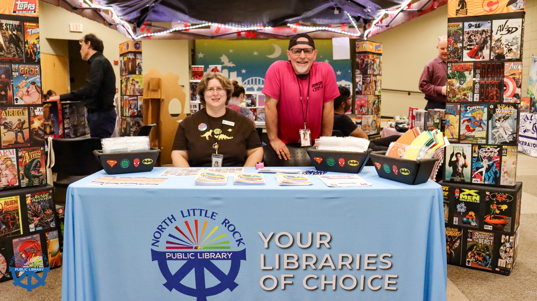 Two people standing behind a table with a North Little Rock Public Library banner surrounded by comic book and game booths.