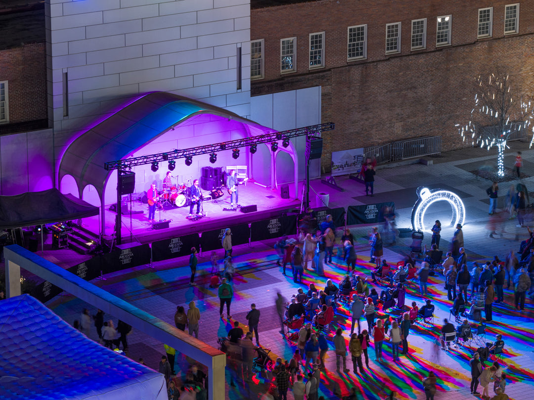 A band playing at an outdoor venue with neon lighting in front of a crowd.