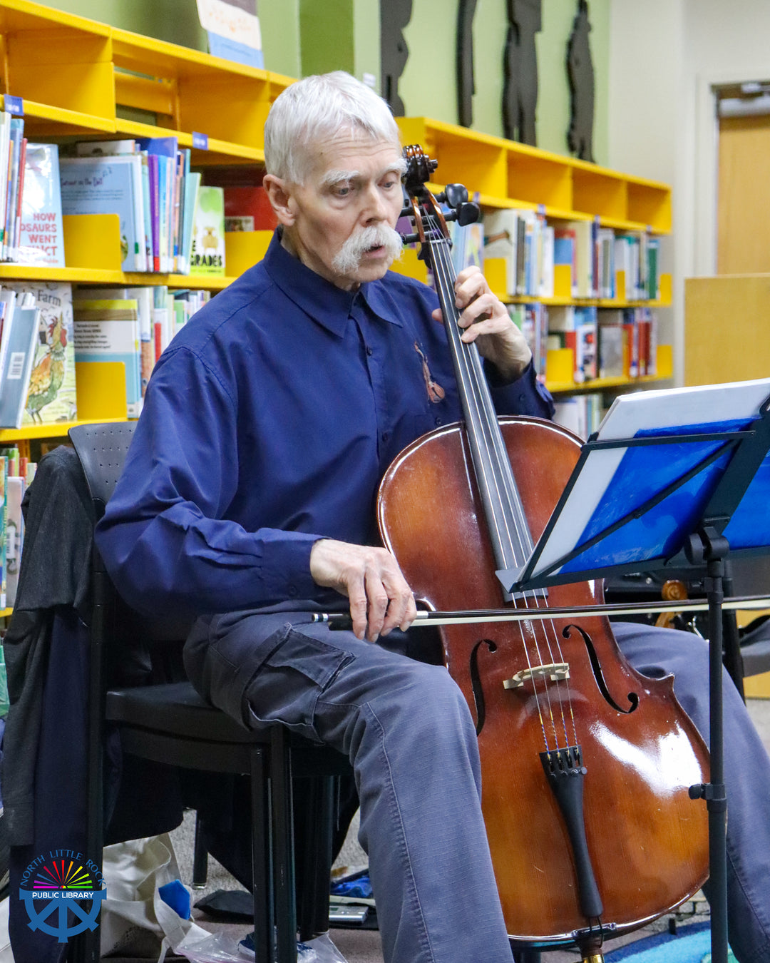 A man with white hair sits with a cello playing music.