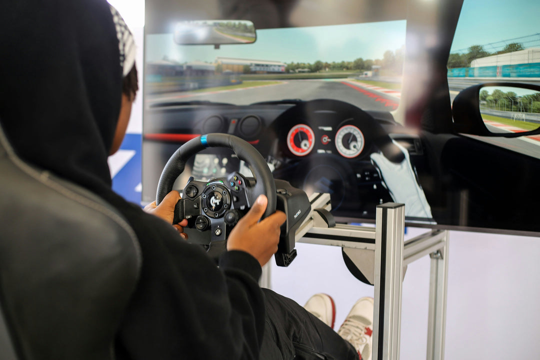 A seated person in a black hoodie with hands on a small car wheel in front of a screen simulating a driving course.