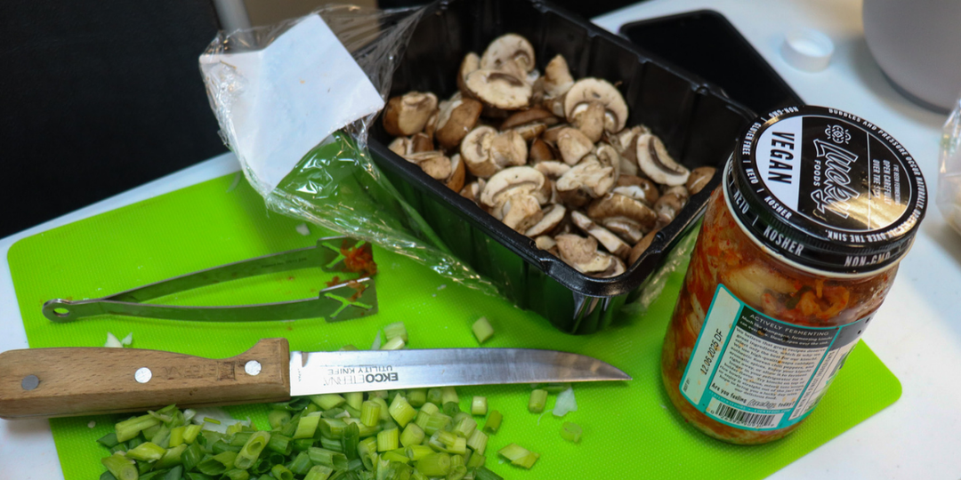 A knife, tongs, chopped green onions, a carton of mushrooms, and a jar of kimchi sitting on a green cutting board.