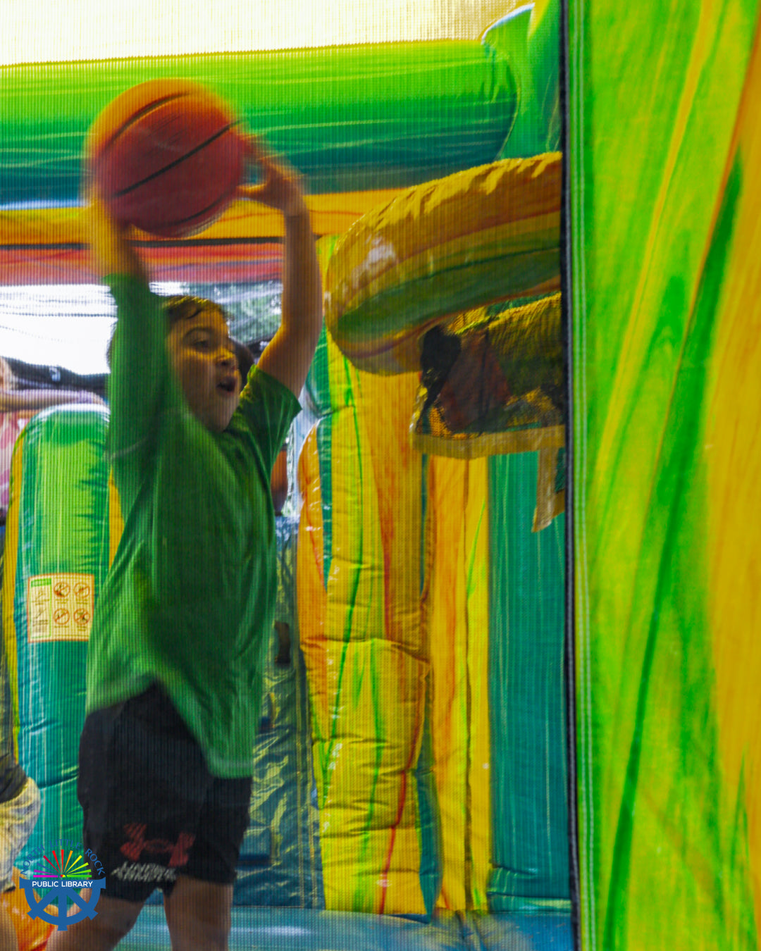 A child prepares to dunk a basketball in an inflated hoop. The photo is taken through the netting of an inflatable water slide.