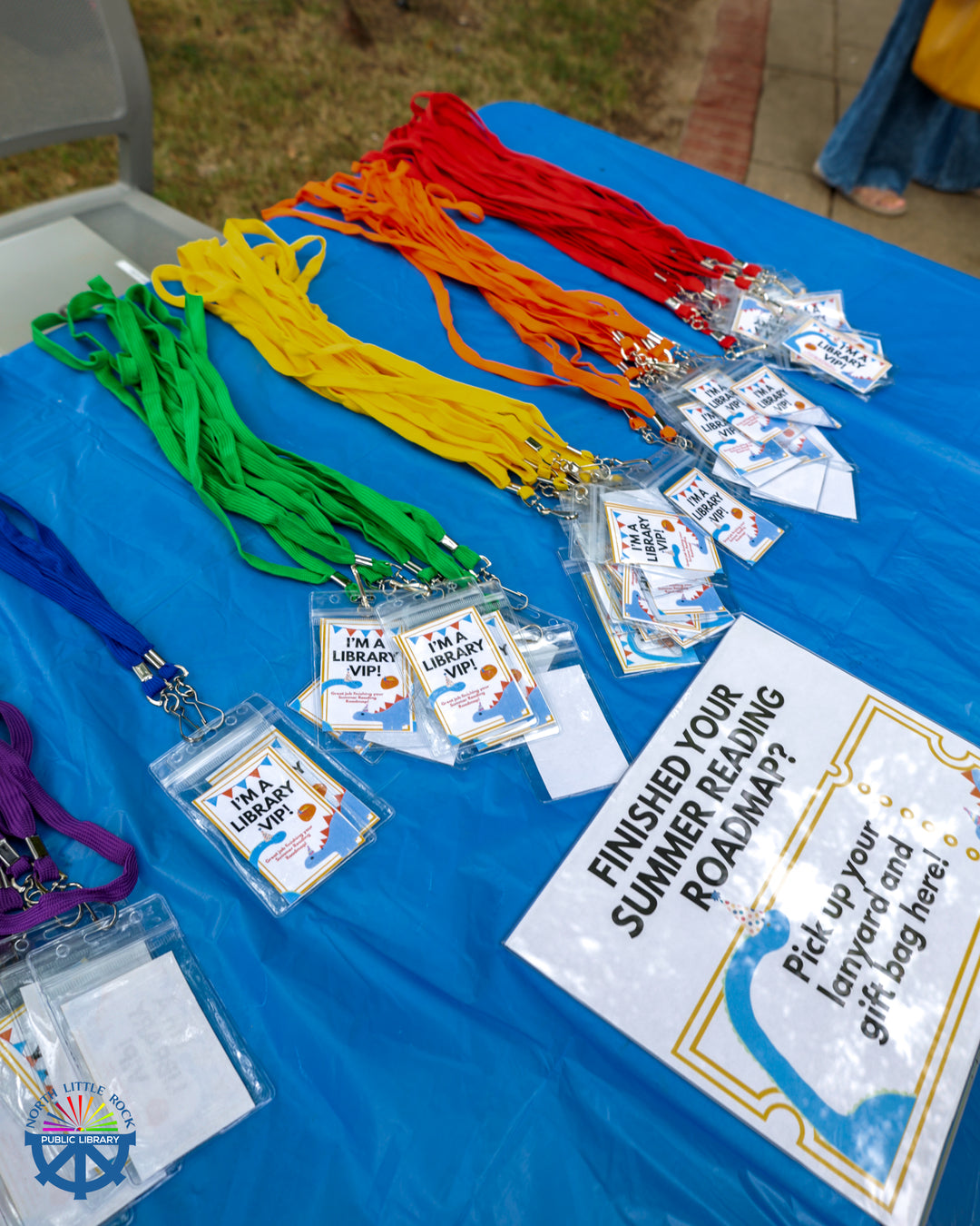 A table covered in a blue tablecloth with colorful lanyards which read I'm a library VIP! In front of the lanyards is a sign reading Finished your Summer Reading Roadmap? Pick up your lanyard and gift bag here.