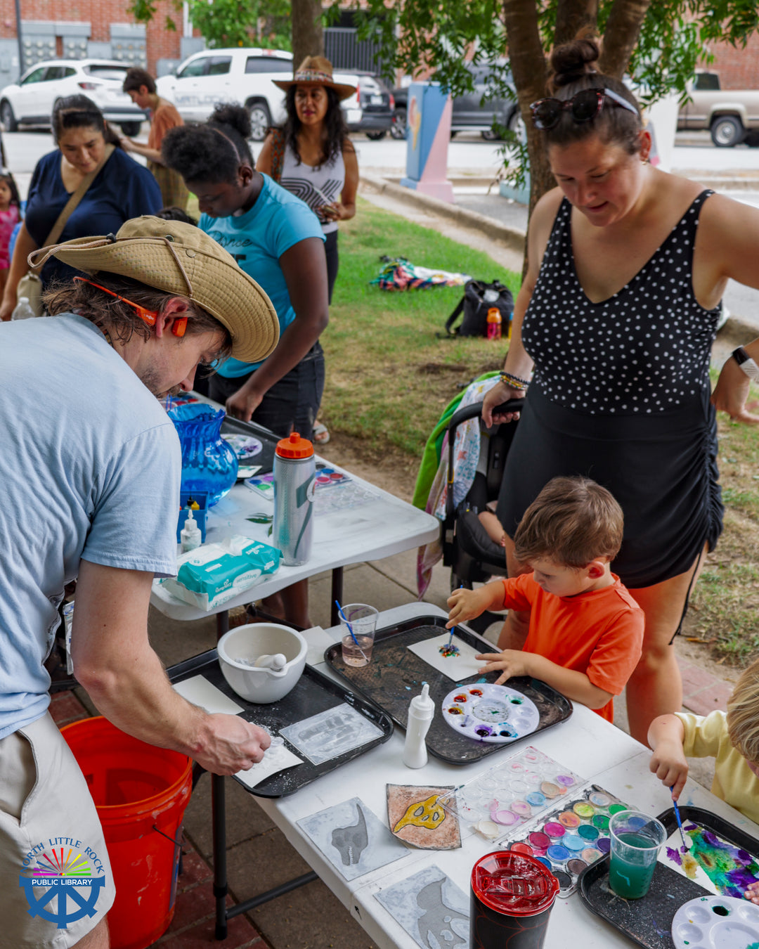 Aaron from the Mobile Makerspace leads families through a dinosaur water color activity. People of all ages stand at white tables, painting and supervising children.