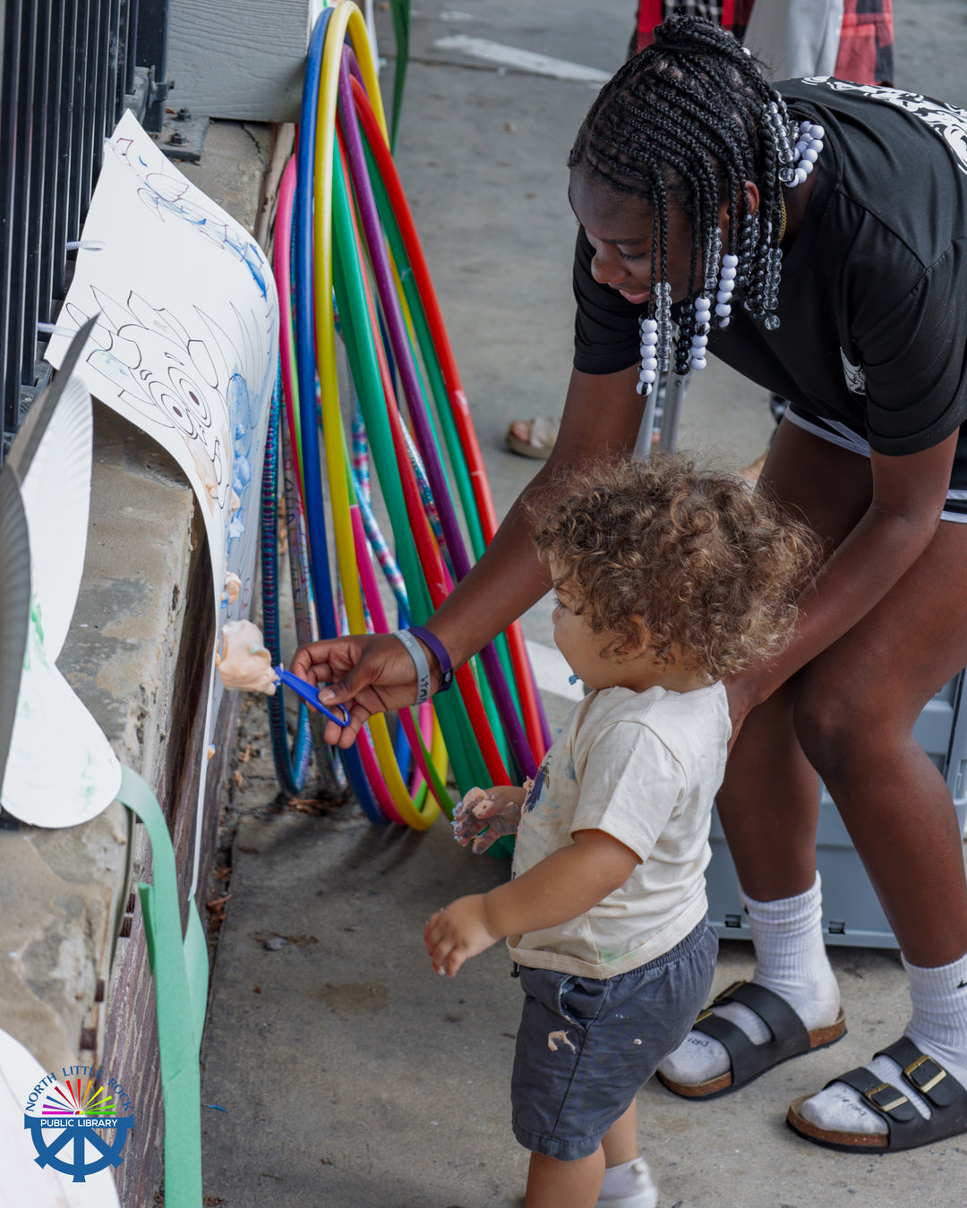 An adult shows a child how to paint using colorful shaving cream. In the background are colorful hula hoops.