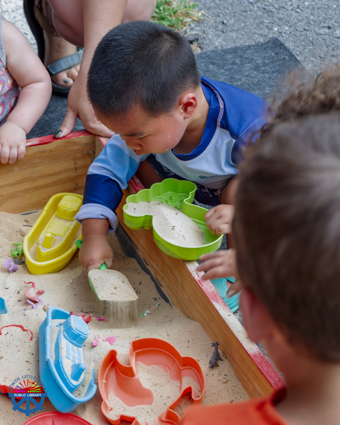 A young child plays in a sandbox with colorful toys and shovels.