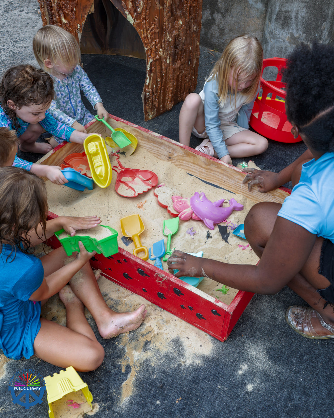 A group of children sit around a red box filled with sand. They play with colorful shovels and toys in the sand.