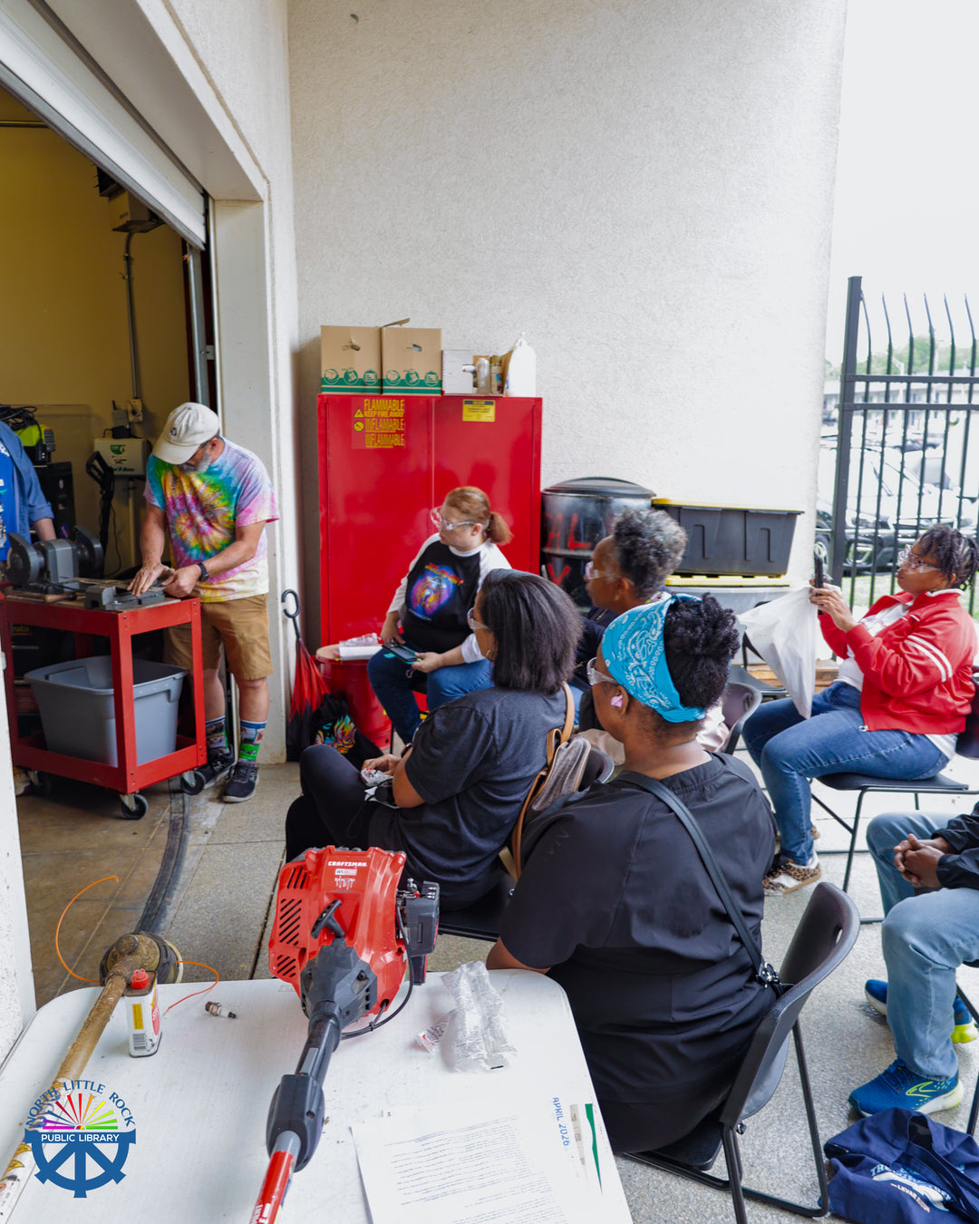 People sitting outside watch a library staff member use a tool to sharpen landscaping equipment. 