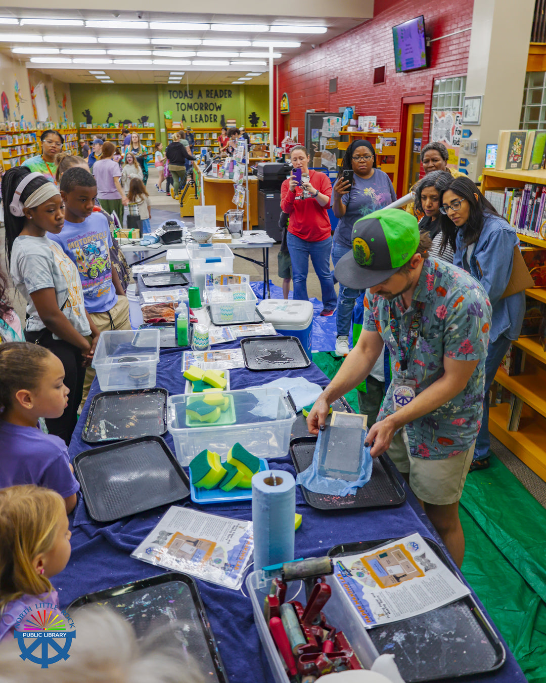 Mobile Makerspace supervisor Aaron demonstrates papermaking to a group of people. They stand in front of tarp-covered tables, lined with trays, spongest, containers, paper towels, and more.