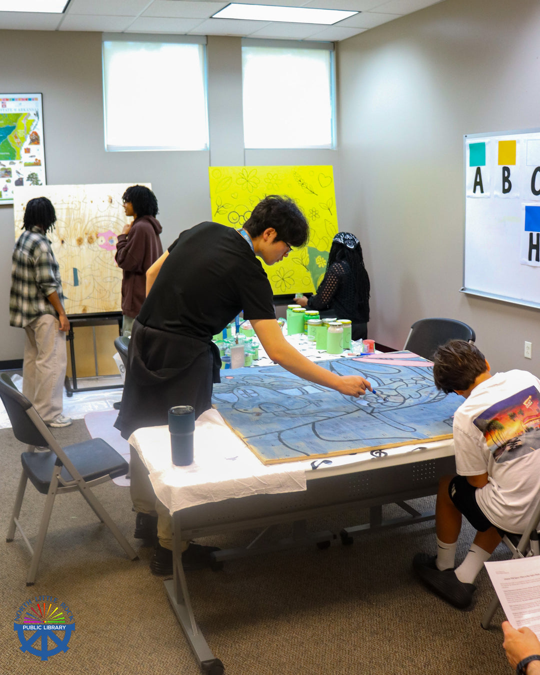 A group of people paint mural panels in the library's Room 124 with a variety of colors and designs.
