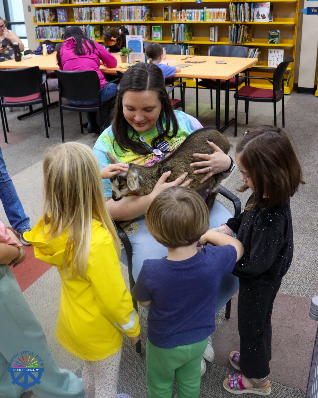 Laman children's department manager Jill holds a baby goat on her lap. A group of children stands around, ready to pet the goat.