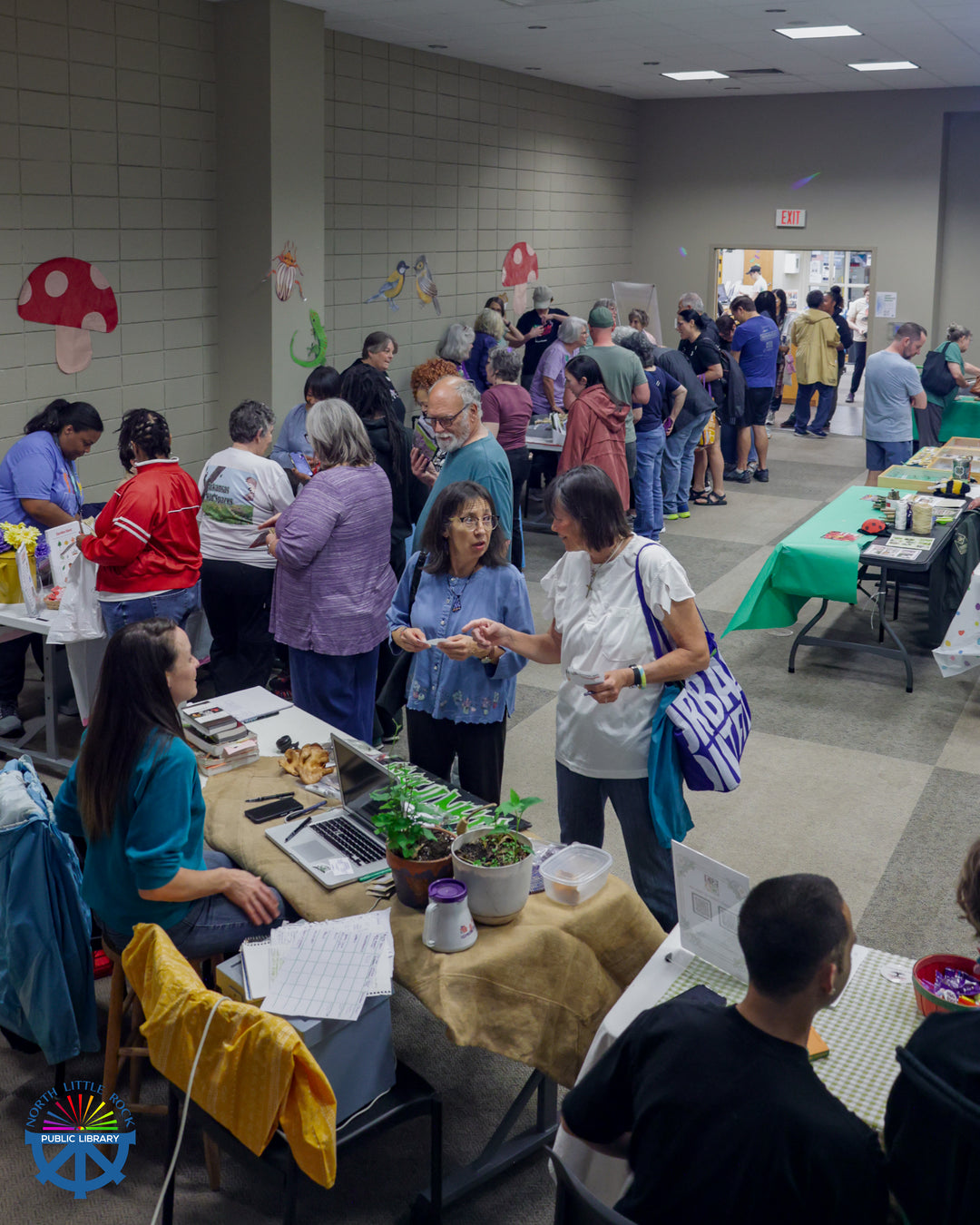 Attendees move from table to table in the library's Lecture Hall, learning about local businessnes and organizations. The room is decorated with colorful birds, bugs, and mushrooms.