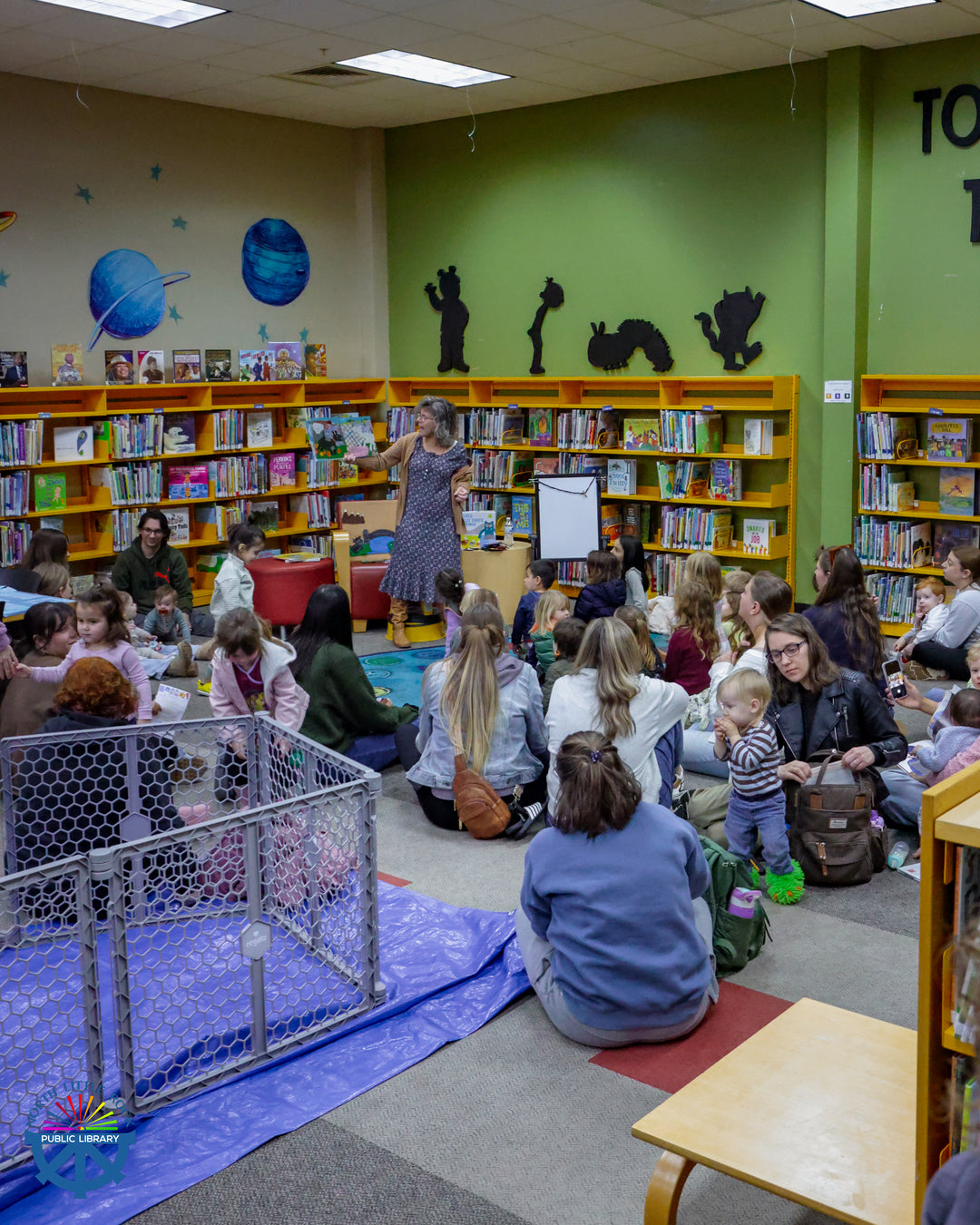 Laman children's staff member Dawn leads a goat-themed storytime in colorful room with bookshelves for seated children and caregivers.
