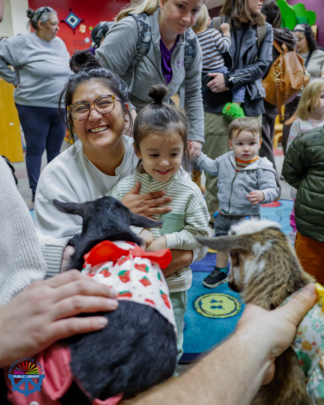 An adult and child smile and two baby goats wearing dresses. A crowd of people wait in line behind them to see the goats.
