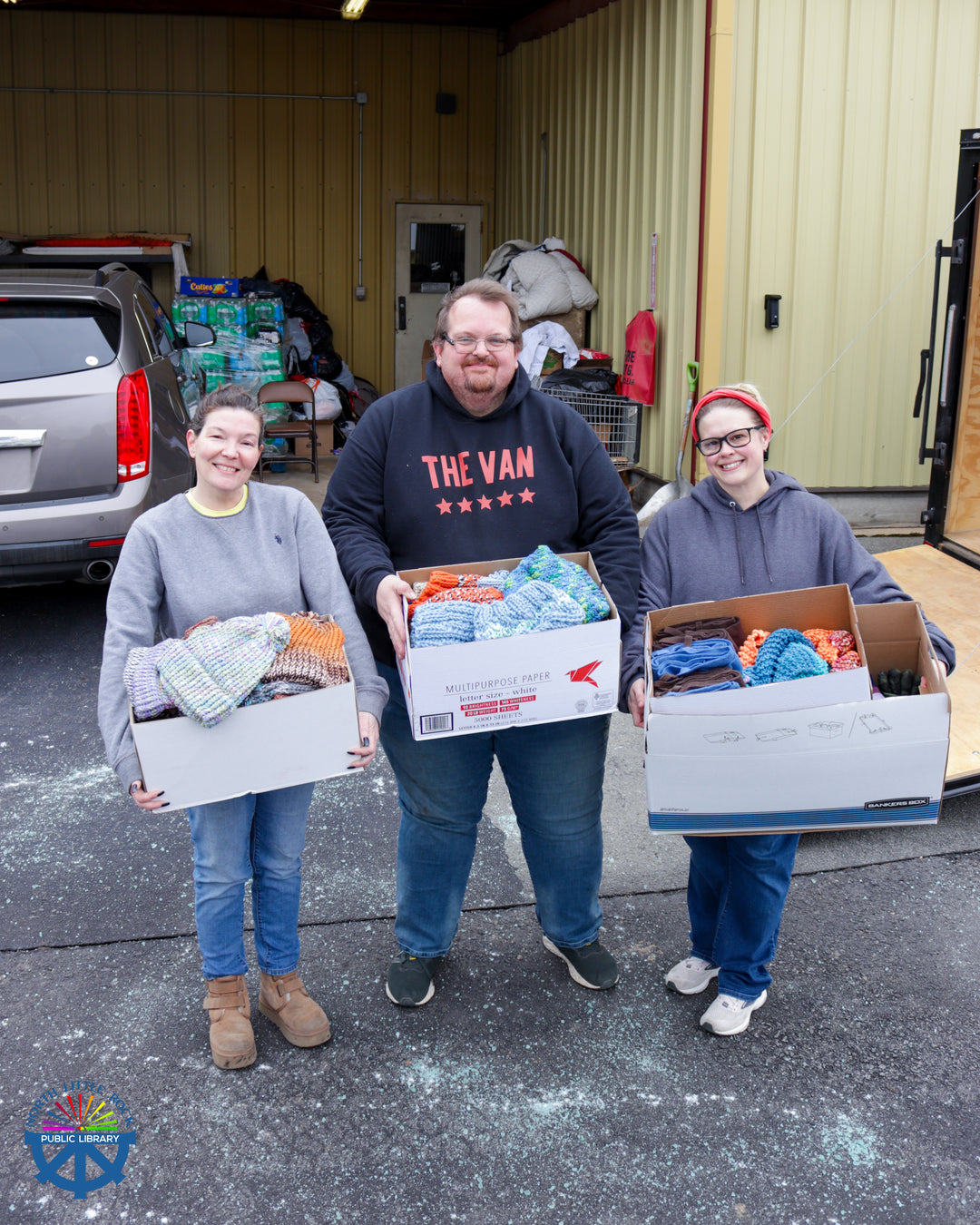 Three people holding boxes full of warm hats from the library.