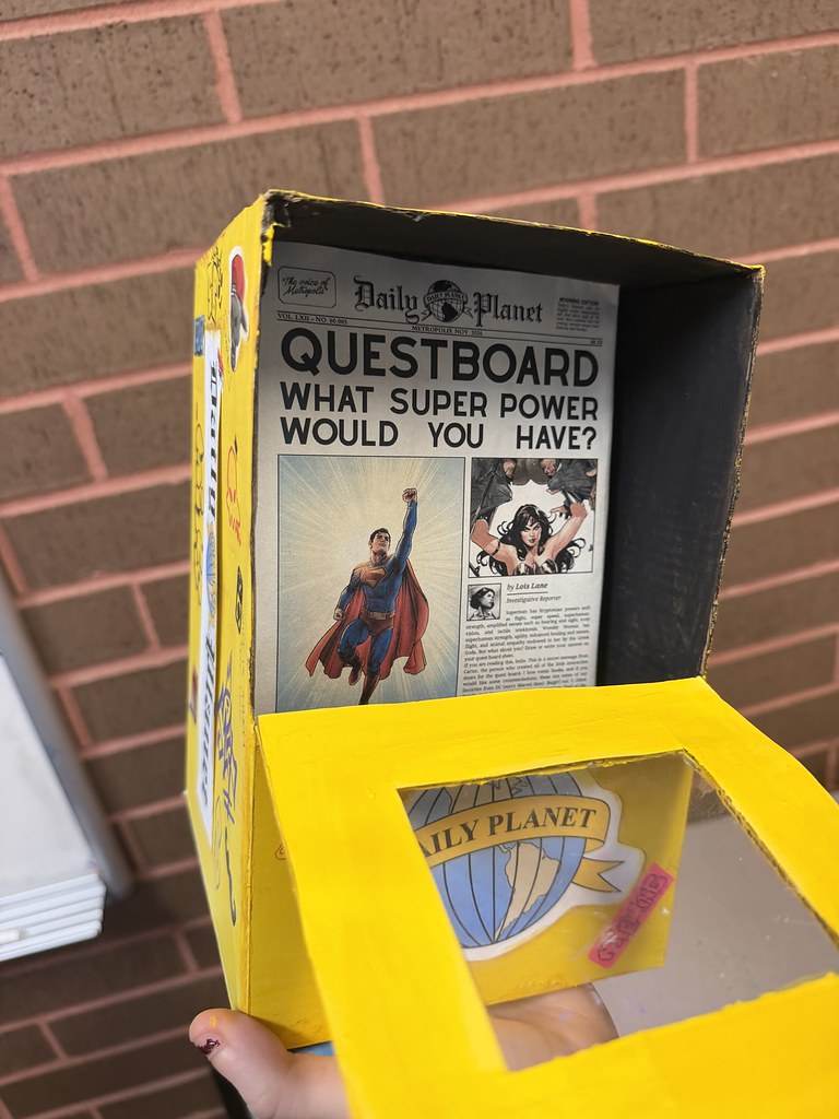 A cardboard box painted yellow and stylized to look like a comic book. Inside is a page of a newspaper reading Questboard: What superpower would you have?
