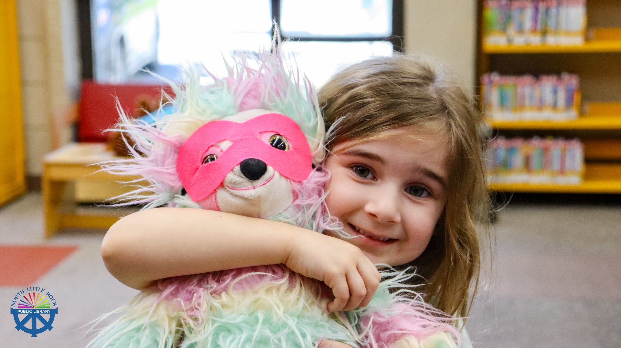 Child holding a colorful teddy bear with a pink superhero cosplay mask in north little rock library setting