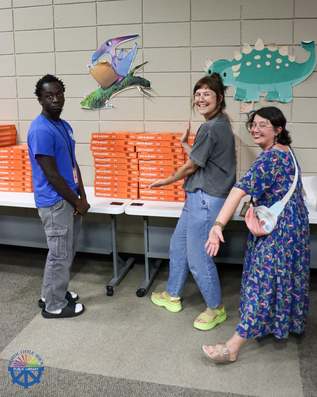 Three library staff members show off tall piles of donated pizza boxes on tables.