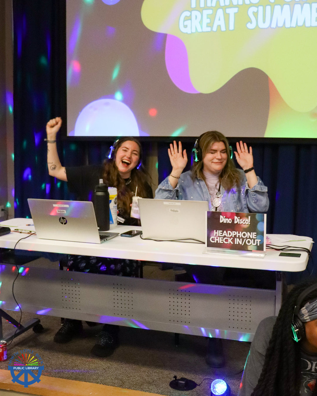 Two library staff members sitting at a table with laptops and headphones playing music for the Silent Disco.