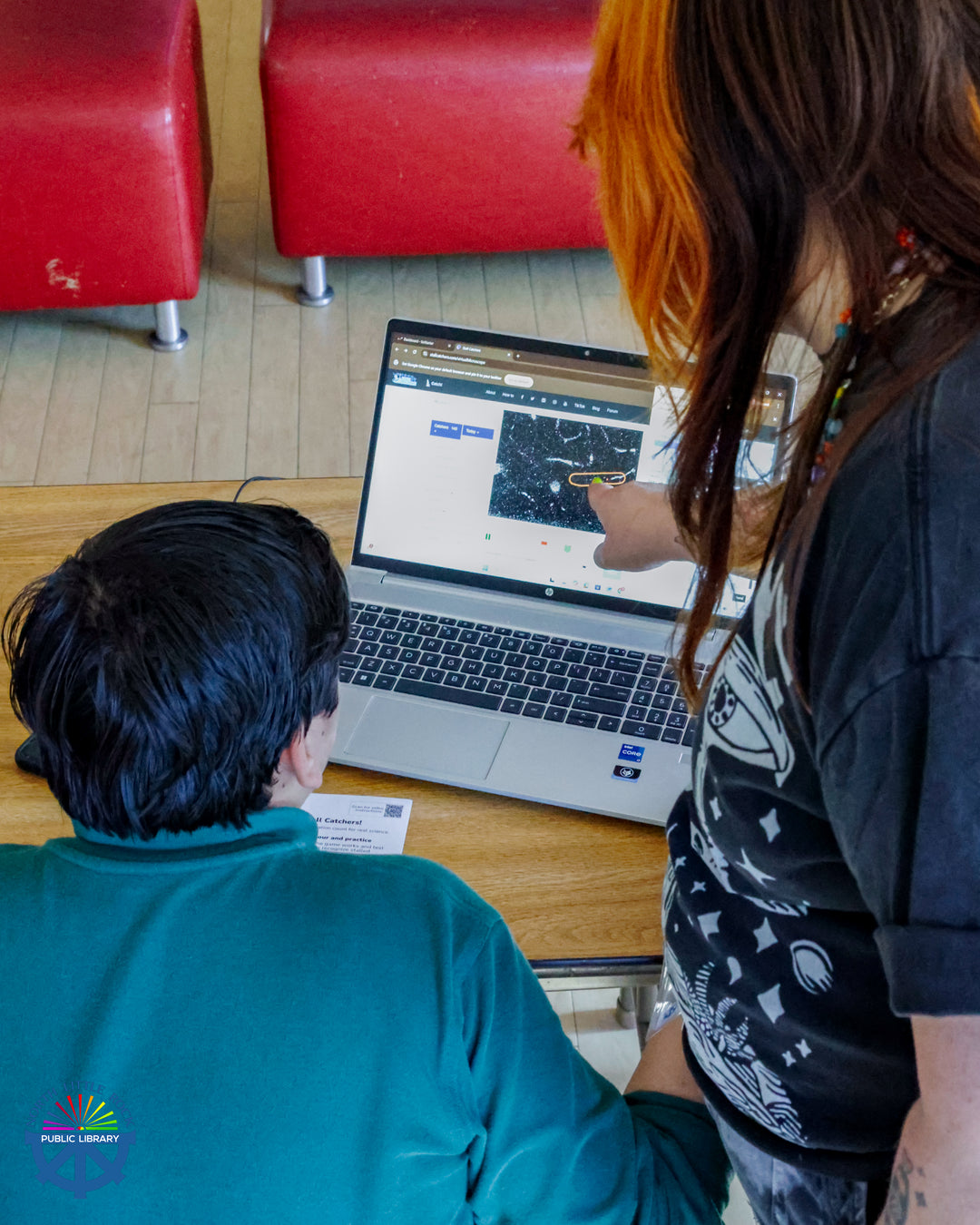 A person sits in front of a laptop while a standing person points at the screen.