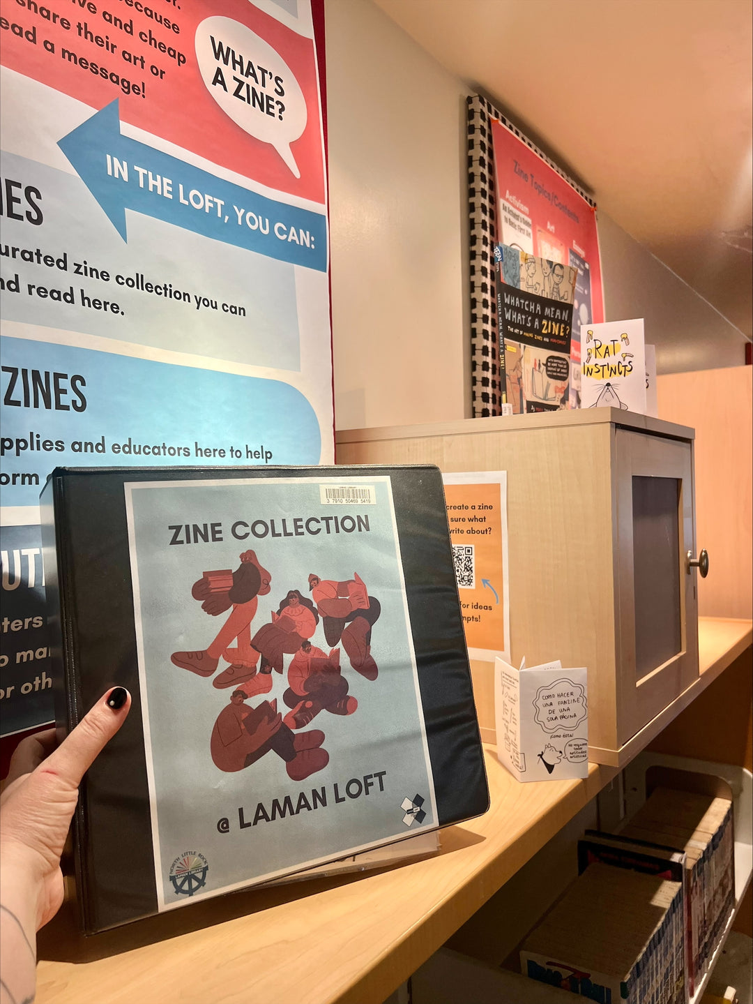 A binder reading "Zine Collection at Laman Loft" in front of a cabinet and several posters about zines.