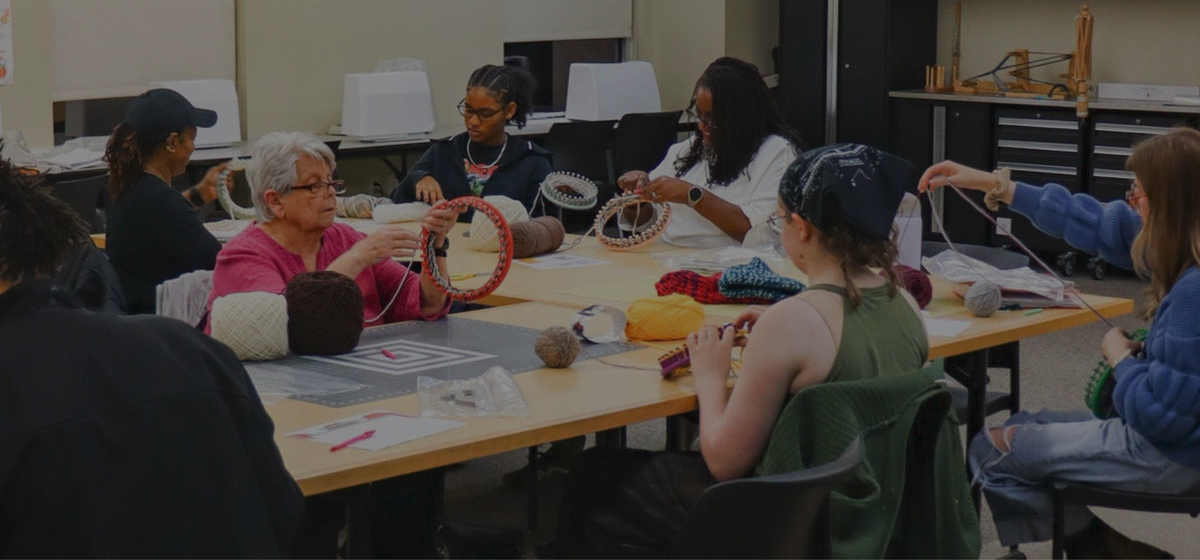 A group of of people sit at a table, knitting yarn hats on colorful looms.