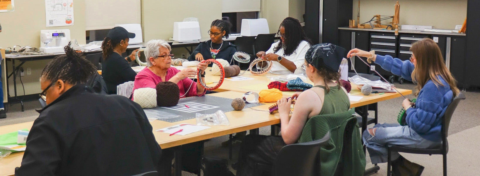 A group of people sitting at tables using looms to knit hats.