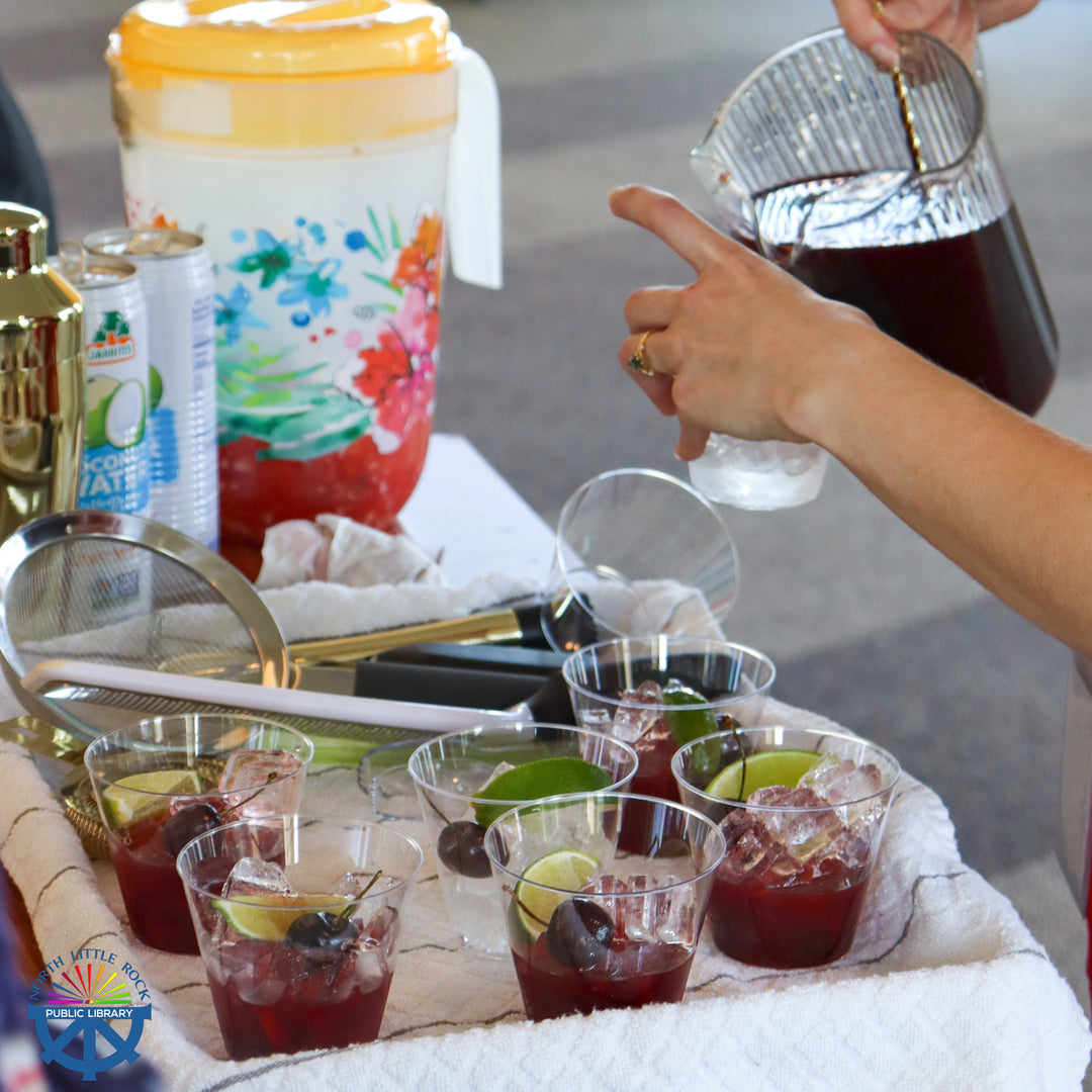 A tray of cups filled with a cherry mocktail with various garnishes. A person fills a cup from a pitcher.