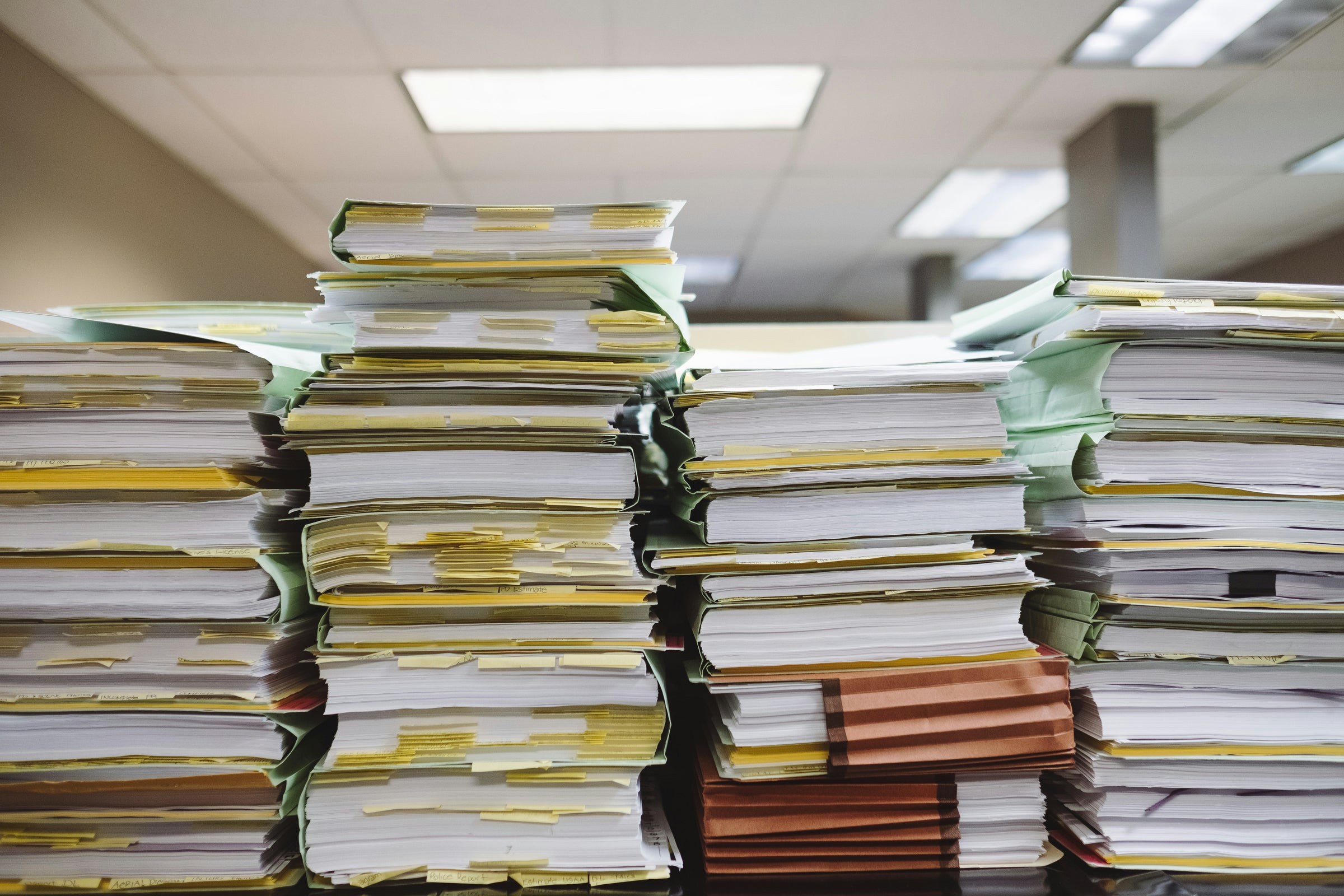 Piles of documents in folders with sticky notes in an office setting.