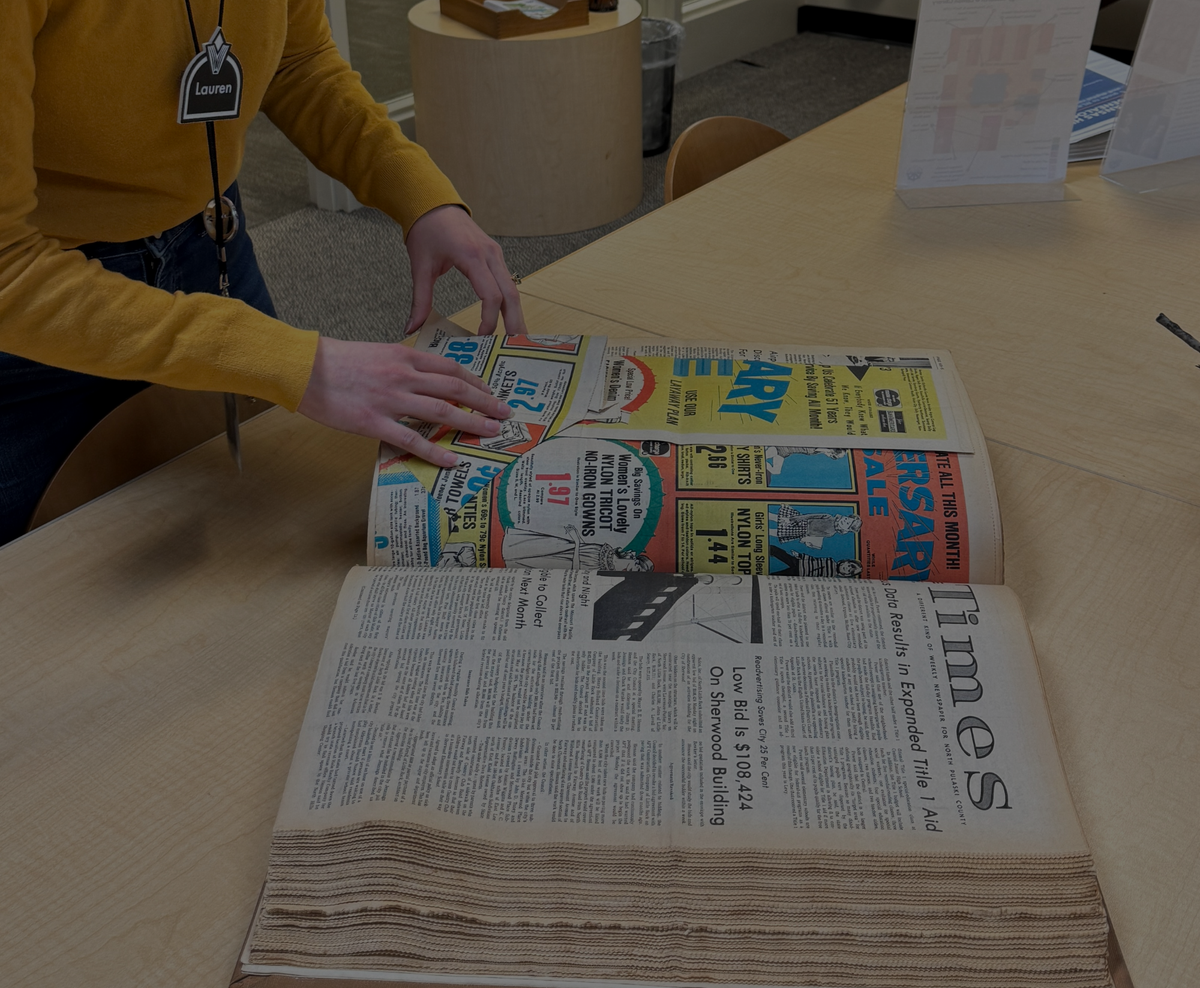 A library staff member looks at a book of newspapers on a wooden table.