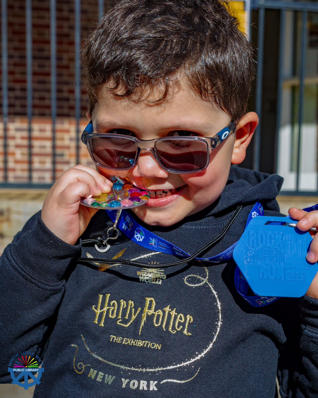 A child in a sweatshirt and sunglasses bites a colorful 5k medal.