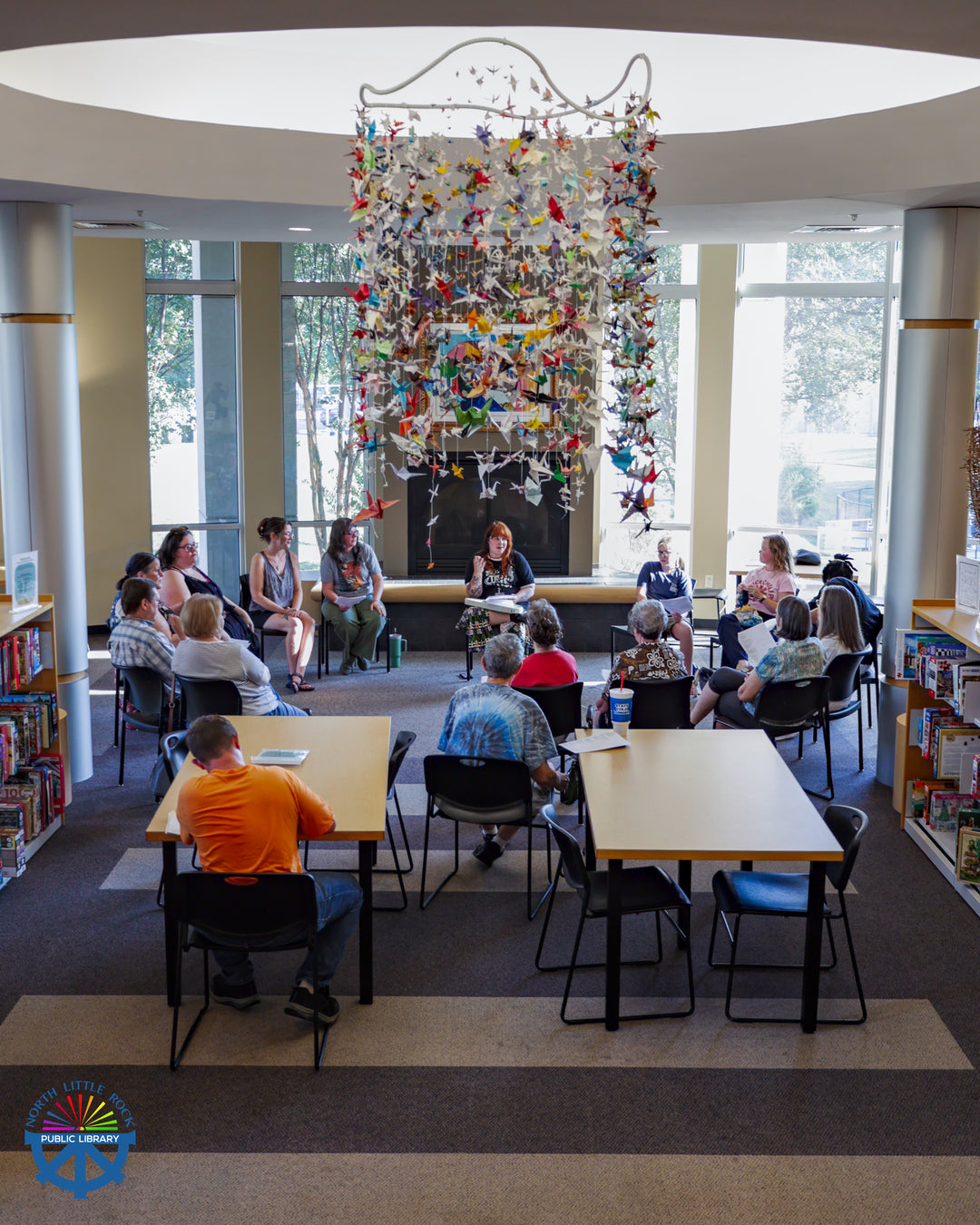 Library staff member Jenna leads a group of seated people in a shared song. Above them hangs an art piece made of hundreds of colorful paper cranes.
