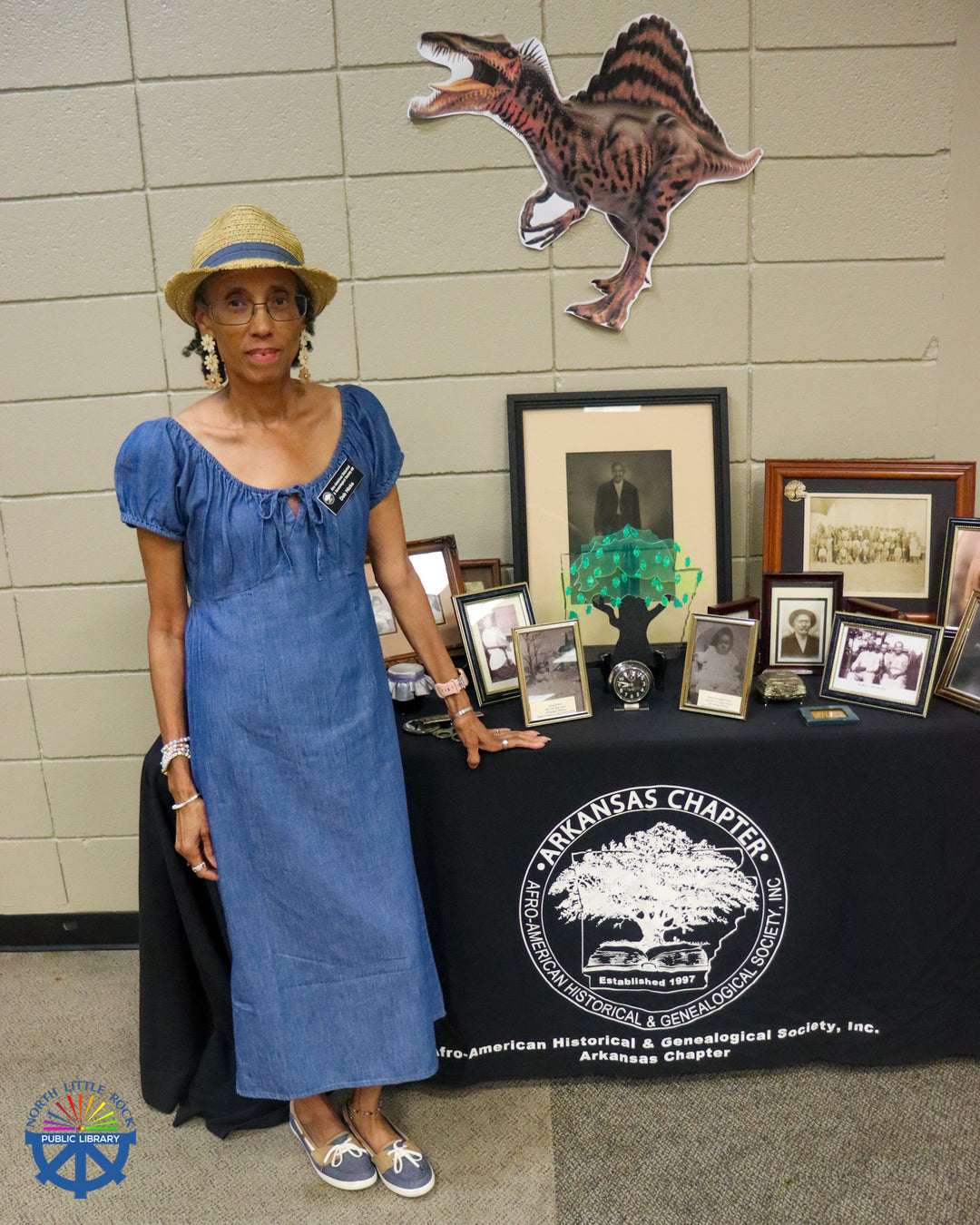 A presenter stands next to photographs and memorabilia on a table with a tablecloth reading Afro-American Historical and Genealogical Society, Inc. Arkansas Chapter.