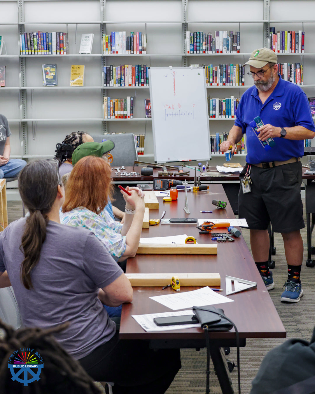 Argenta maintenance person Kendell demonstrates hand tool basics at a Mr. Fix-it workshop. Attendees sit at wooden tables with a variety of wood blocks and tools while Kendell demonstrates.