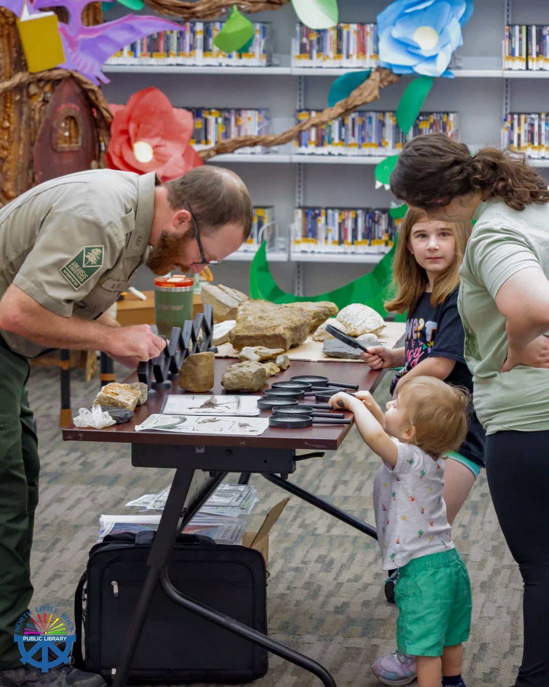 Parks Interpreter Matthew stands in front of a table with rocks and tools, explaining what they are to a family on the other side of the table.