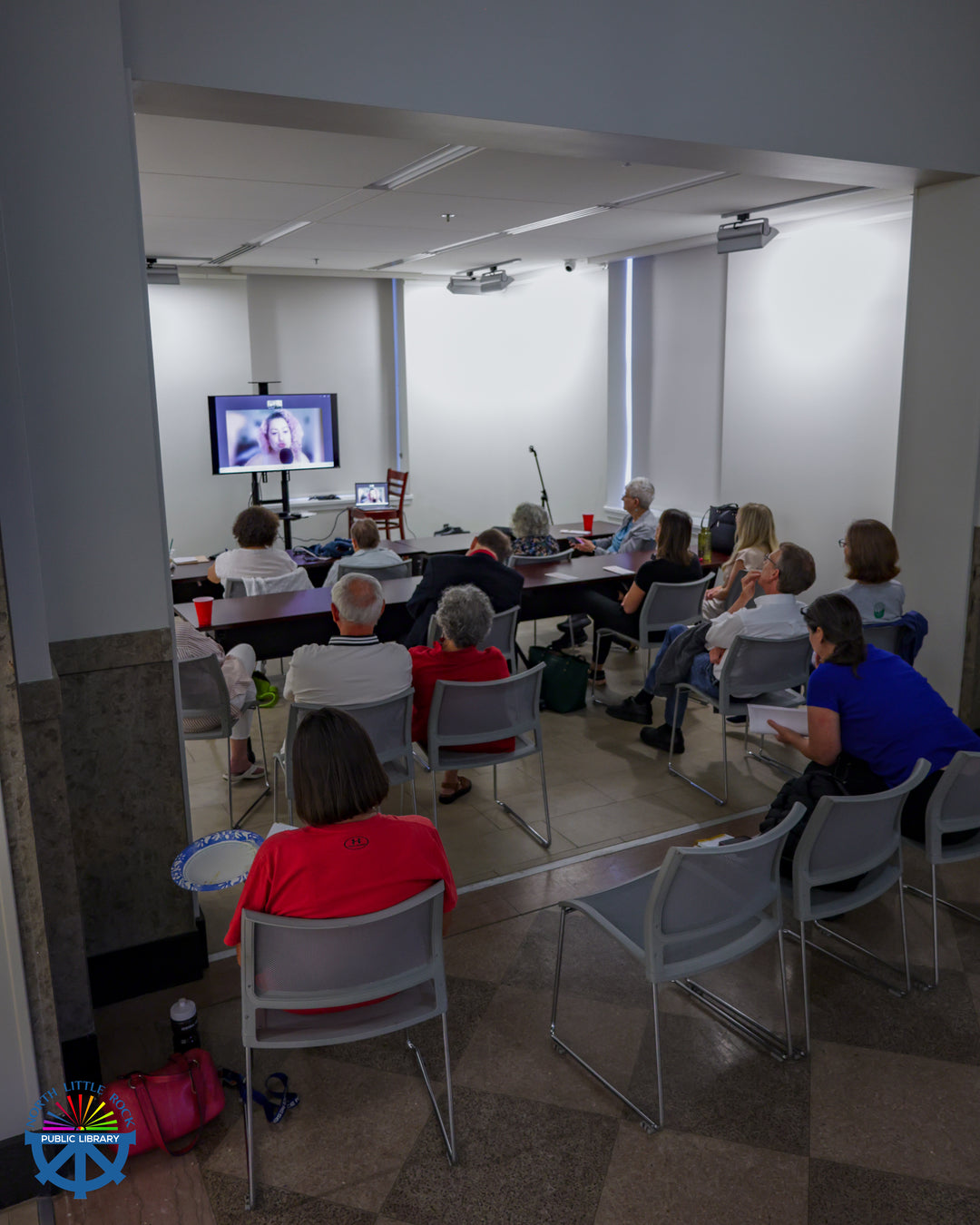 A group of seated people watch a woman on a TV screen talk about Yiddish folk traditions and magic. 