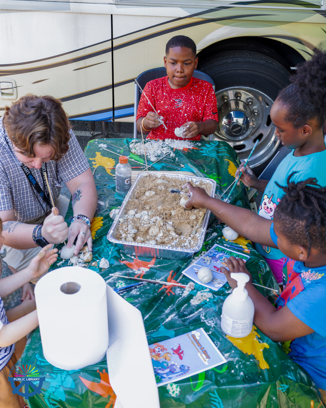Children sit around a table with Rover manager Annika excavating dinosaurs with skewers.