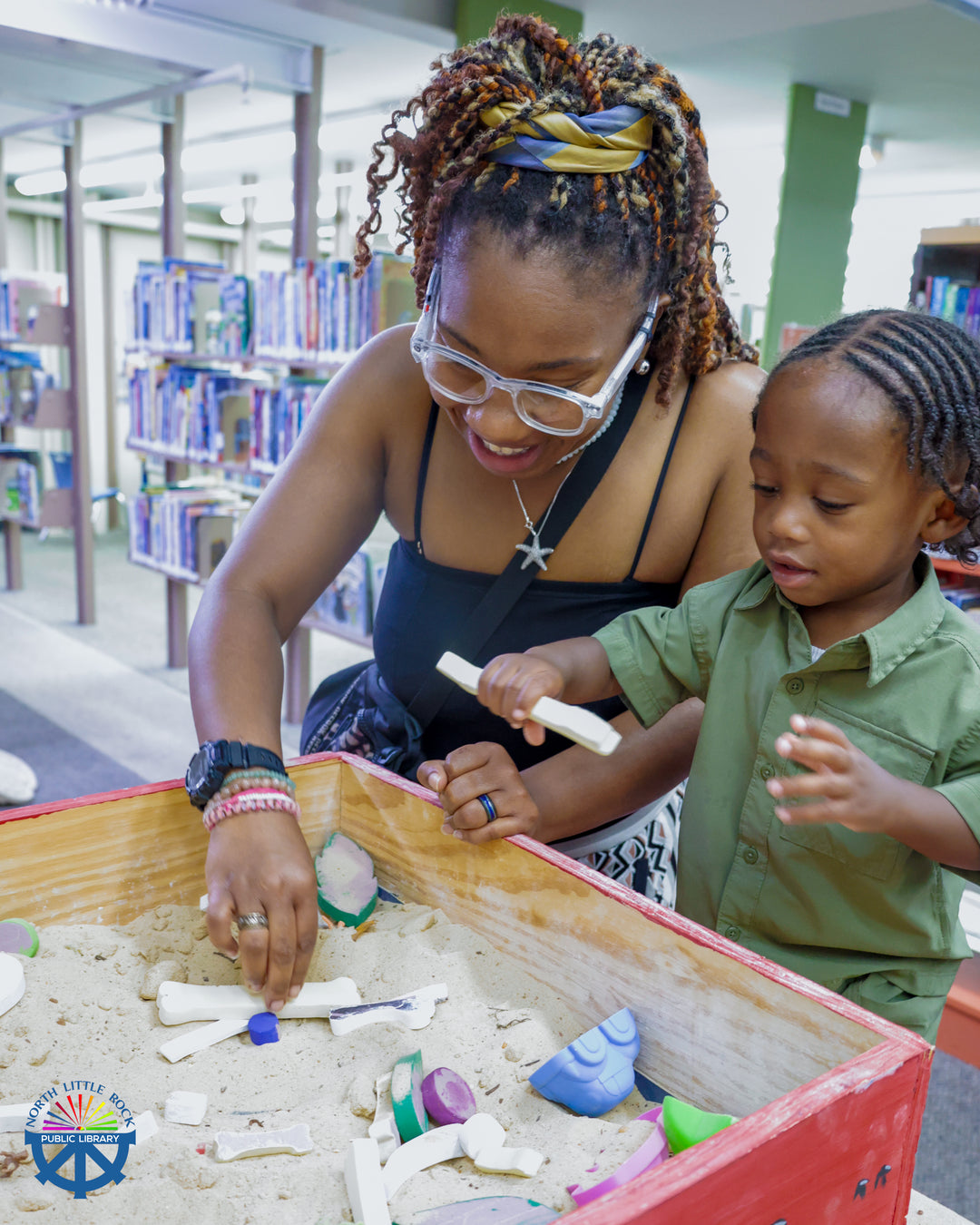 An adult and child play in a sandbox, excavating toy dinosaur bones.